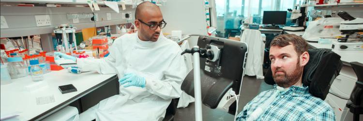 A researcher in a lab coat is sitting at a laboratory bench, talking through laboratory procedures with Euan Macdonald who is in his power-chair, Euan is engaging the researcher in conversation 