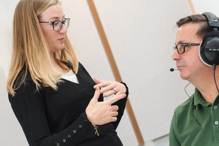 A SpeakUnique researcher is talking with a participant, who is wearing with headphones with a microphone piece. The researcher is describing the process for voice banking