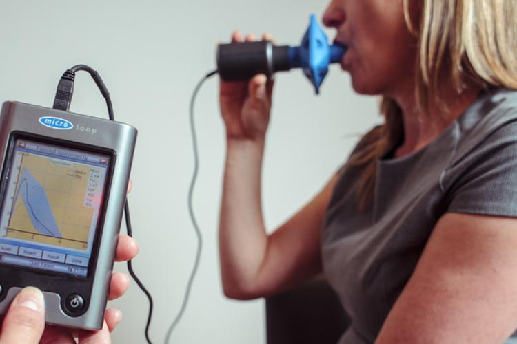 A close up of the screen of a spirometer (an instrument that measures breathing), with an out-of-focus patient in the background doing the test, by blowing into the mouthpiece