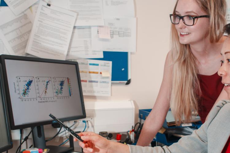 Two PhD students are looking at two computer screens with graphs on one, brain slice scans on the other
