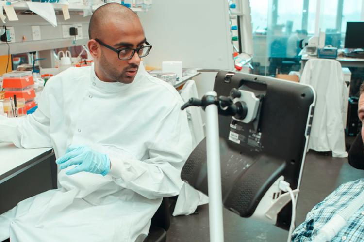 A researcher in a lab coat is sitting at a laboratory bench, talking through laboratory procedures with Euan Macdonald who is in his power-chair, Euan is engaging the researcher in conversation 