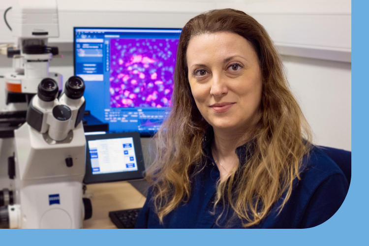 A picture of Ilary Allodi in her laboratory with a microscope in the background showing images of cells brightly coloured purple