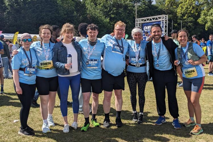 Eight people standing with linked arms outside near the finish line of the Edinburgh marathon relay, wearing light blue Euan MacDonald Centre T-shirts and with medals around their necks