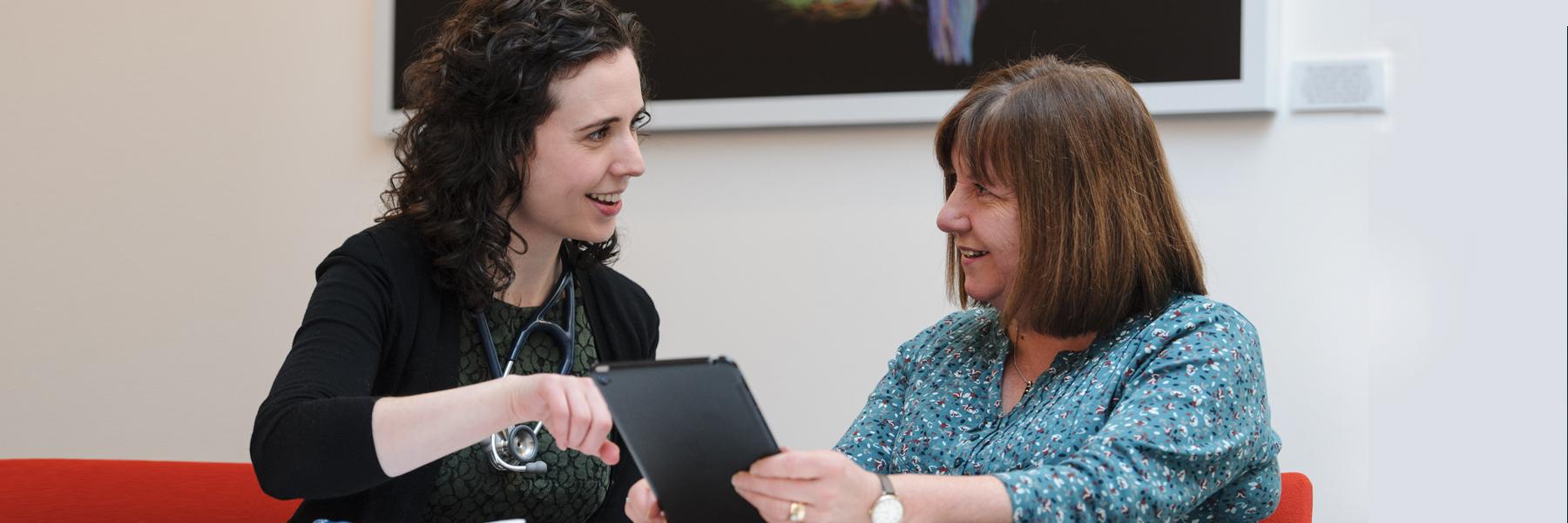 Doctor and patient picture happily talking while patient holds an iPad and Doctor is pointing at the screen