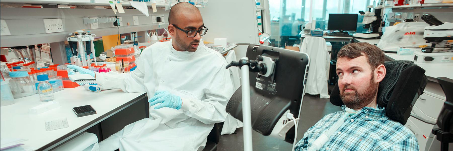 A researcher in a lab coat is sitting at a laboratory bench, talking through laboratory procedures with Euan Macdonald who is in his power-chair, Euan is engaging the researcher in conversation 