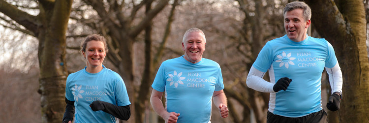 Three fundraisers in a park wearing blue Euan MacDonald Centre t-shirts they are running and smiling at the camera