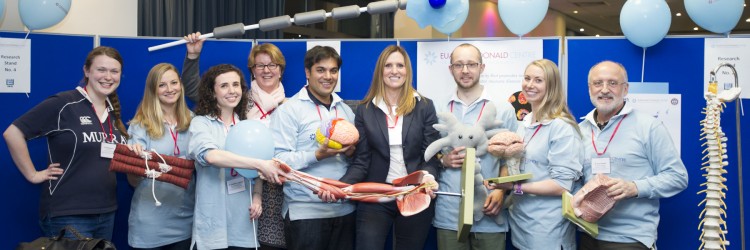 a group of nine Euan MacDonald Centre members at an event, holding props they have used to demonstrate their research