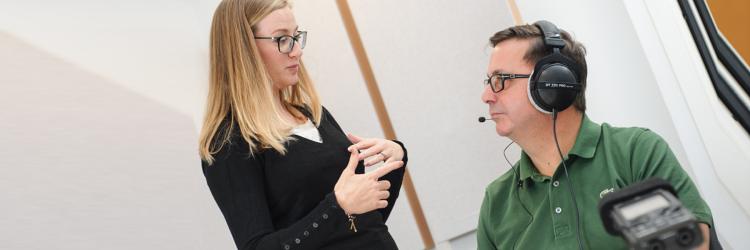A researcher is standing talking to a research participant, who is sitting down wearing over the head headphones with a microphone attachment ready to bank his voice