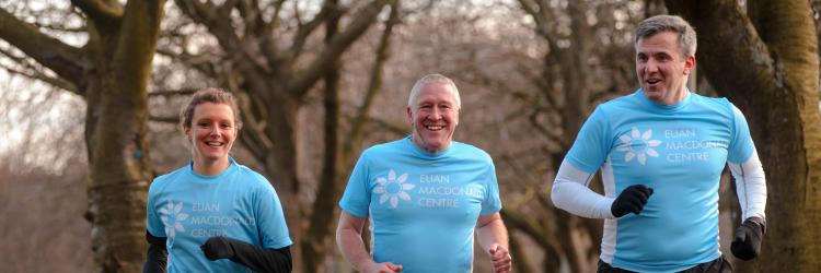 Three fundraisers in a park wearing blue Euan MacDonald Centre t-shirts they are running and smiling at the camera