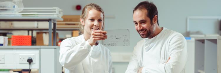 Two PhD students in lab coats smiling and looking at some lab results 
